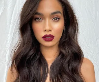 Woman with long, wavy dark hair and bold red lipstick against a light background.