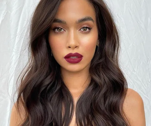 Woman with long, wavy dark hair and bold red lipstick against a light background.