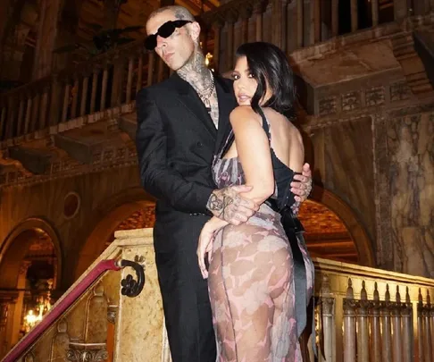 A couple poses on a staircase inside an ornate building, with the man wearing sunglasses and tattoos visible.