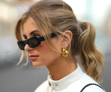 Woman with sunglasses and cartilage earrings, wearing a white top, hair in a ponytail.