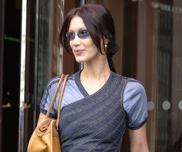 Woman in layered denim outfit with sunglasses and brown handbag walking outdoors.