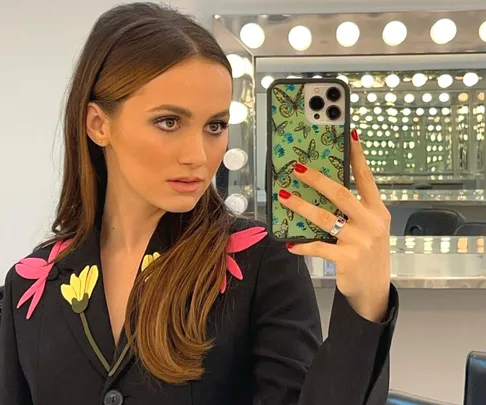 A woman with long brown hair takes a mirror selfie in a floral jacket, in a brightly lit makeup room.