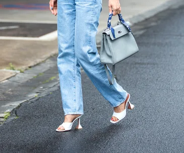 Person walking on wet pavement in blue jeans and white heeled sandals, carrying a gray handbag with a patterned scarf.