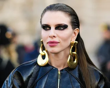 Woman with bold black eye makeup, pink lipstick, large gold earrings, and slicked-back hair wearing a shiny black jacket.