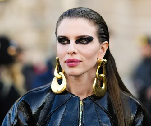 Woman with bold black eye makeup, pink lipstick, large gold earrings, and slicked-back hair wearing a shiny black jacket.