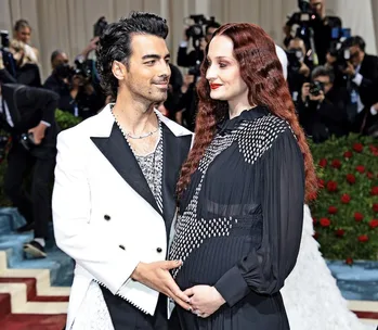 A couple poses on the red carpet, the woman wearing a black dress and displaying a baby bump, while photographers capture the moment.