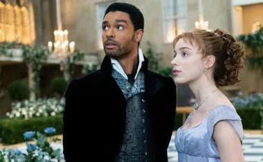 A couple in 19th century attire standing in a lavish ballroom surrounded by flowers and candlelight.