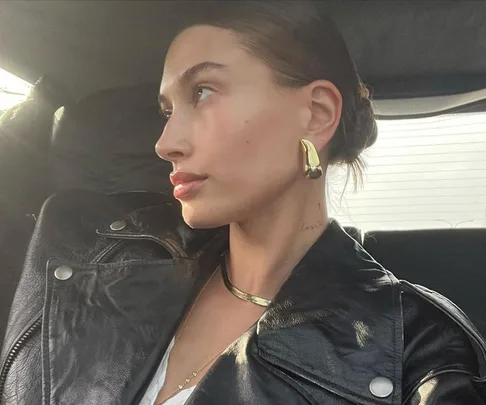 Woman in a black leather coat with gold earrings and a sleek hairstyle, looking out a car window.