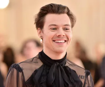 Smiling man in a sheer, black outfit with a pearl earring, standing at an event.