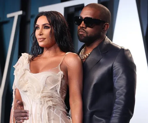 A man in sunglasses and leather jacket poses with a woman in a white dress, both standing in front of a decorative backdrop.