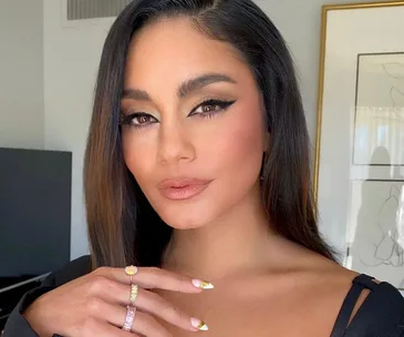 A woman with long dark hair showcases dramatic winged eyeliner, wearing a black top and colorful rings, posing indoors.