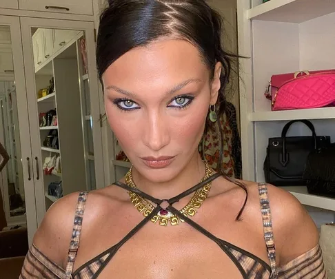 Person with sleek hairstyle and strappy top stands in room with shelves of handbags.