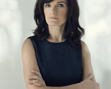 A woman with dark hair and a black sleeveless dress stands with arms crossed against a light background.