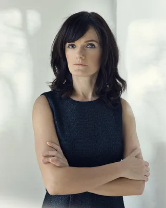 A woman with dark hair and a black sleeveless dress stands with arms crossed against a light background.