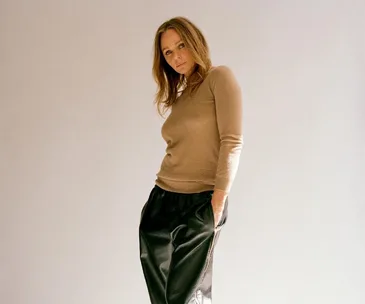 A person posing confidently in a beige top and black trousers with a neutral background.