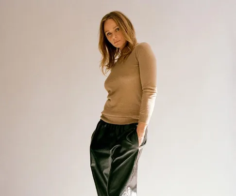 A person posing confidently in a beige top and black trousers with a neutral background.