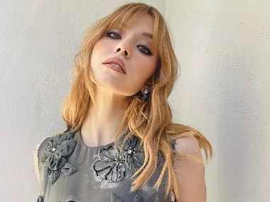 A woman with long blonde hair wearing a floral-patterned dress poses against a pale background.