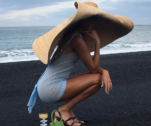 Model crouching on a black sand beach in a large straw hat and light blue dress, with a green bag and drinks nearby.