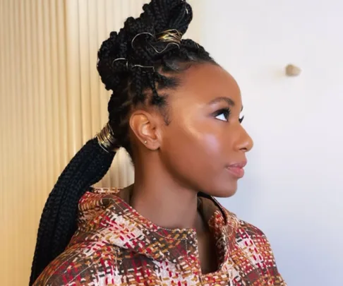 Profile of a person with intricate braided hairstyle and colorful patterned outfit against a neutral background.