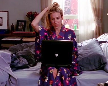 Woman in floral robe using a laptop on bed, pulling hair back, daylight through window. Cozy bedroom setting.