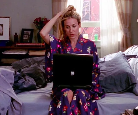 Woman in floral robe using a laptop on bed, pulling hair back, daylight through window. Cozy bedroom setting.