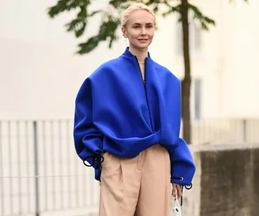 A person in a bold Yves Klein blue oversized jacket with beige trousers, standing outdoors, with trees in the background.