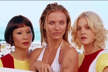 Three women from Charlie's Angels wearing colorful beach attire, standing in front of a sunny beach backdrop.