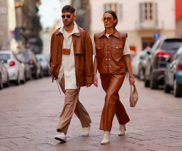 A stylish duo in brown leather outfits, walking down a city street with cars parked along the sides.