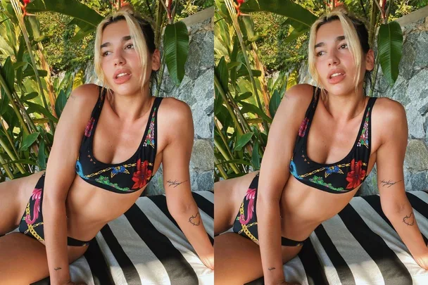 Singer wearing a floral bikini with visible tattoos, sitting outdoors surrounded by plants.