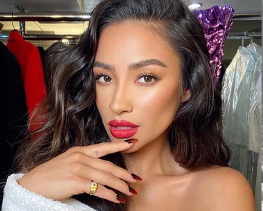 Person with long dark hair and bright red lipstick posing with hand near face in a dressing room setting.