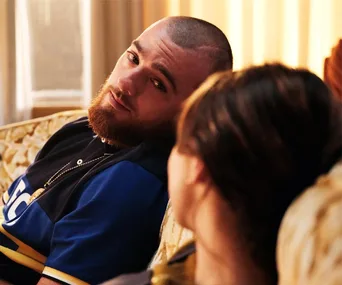 A bearded man in a blue shirt sits on a couch, looking intently at a woman with blurred brown hair.