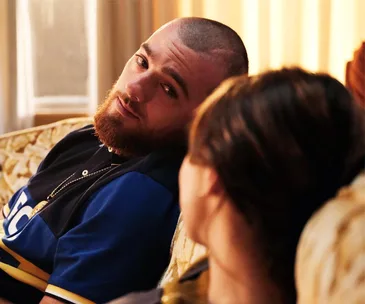 A bearded man in a blue shirt sits on a couch, looking intently at a woman with blurred brown hair.