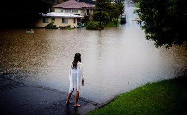 A person stands on a flooded street, observing submerged houses and vehicles in heavy rain.