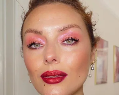 Woman with glittery pink eyeshadow and red lipstick, wearing pearl earrings. Light background with framed art pieces.