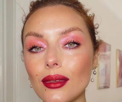 Woman with glittery pink eyeshadow and red lipstick, wearing pearl earrings. Light background with framed art pieces.