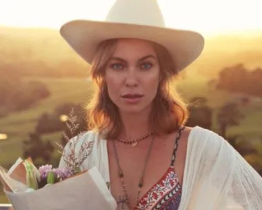 Woman in a hat holding flowers at sunset, standing outside with a scenic countryside background.