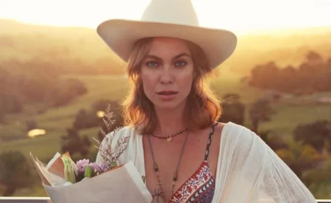 Woman in a hat holding flowers at sunset, standing outside with a scenic countryside background.