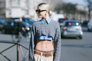 Woman wearing sunglasses, a blue shirt, and a cropped gray sweater, styled with beige pants and a brown belt on the street.