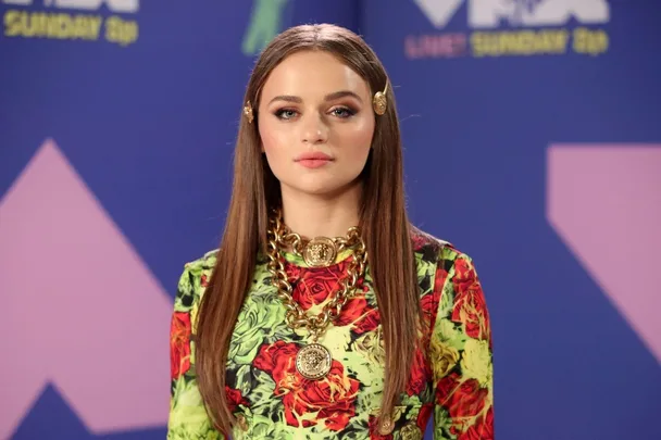 A person in a colorful floral outfit with long hair and gold accessories at an event with a blue background.