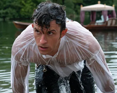 A man in a wet white shirt emerging from a lake, with a boat visible in the background.