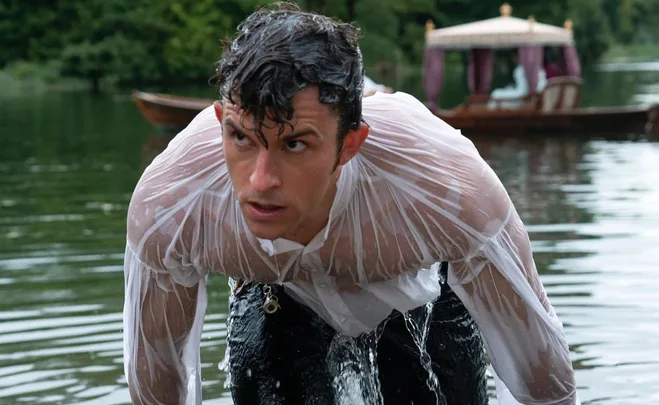 A man in a wet white shirt emerging from a lake, with a boat visible in the background.