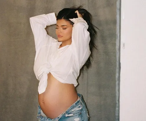 Pregnant woman in a white shirt and jeans, touching her hair, posing sideways with her belly exposed.