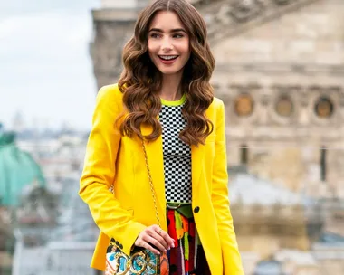 Lily Collins as Emily Cooper, smiling in a vibrant yellow jacket on a city balcony scene from "Emily in Paris."