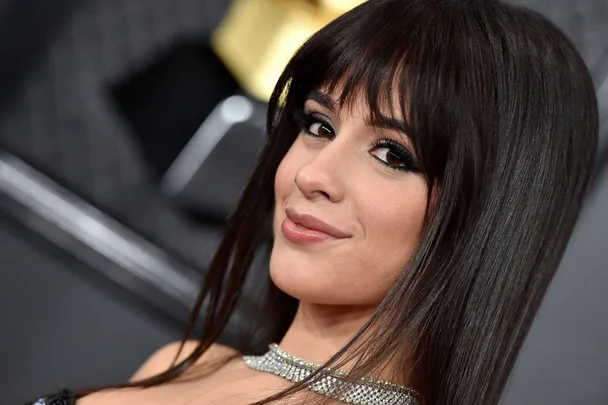 Singer with brown hair and bangs, wearing a sparkly necklace, poses at an event with a confident smile.