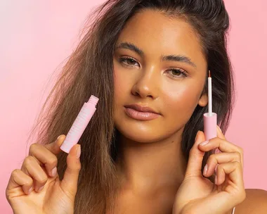 Woman holding Lash Rehab serum against a pink background.