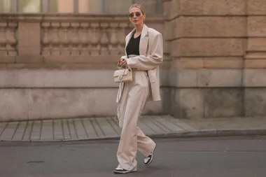 Woman in beige suit and sneakers, carrying a Chanel bag, standing on a city street.