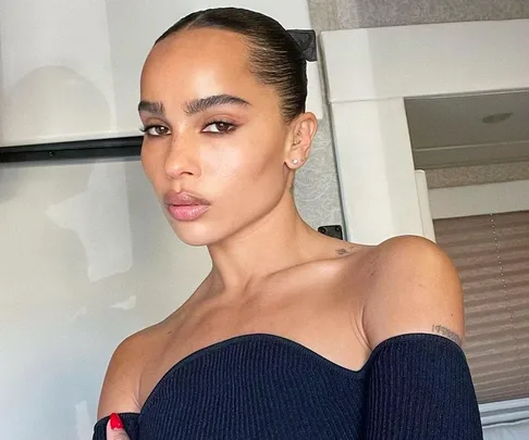 Young woman with slicked-back hair and off-shoulder top, posing indoors with a neutral expression.