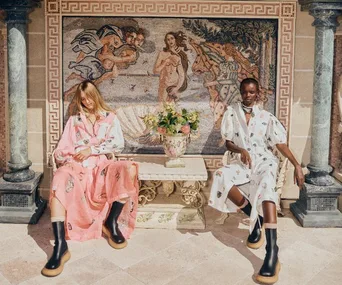 Two women in patterned dresses sit by a decorative mosaic with floral centerpiece; elegant, classical ambiance.