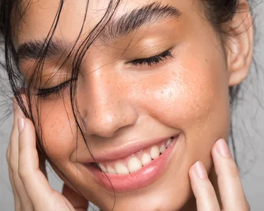 Smiling woman with clear, glowing skin and closed eyes touching her face gently.