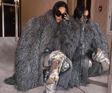 Person wearing a silver textured coat and metallic thigh-high boots crouching inside a hallway with dark sunglasses.